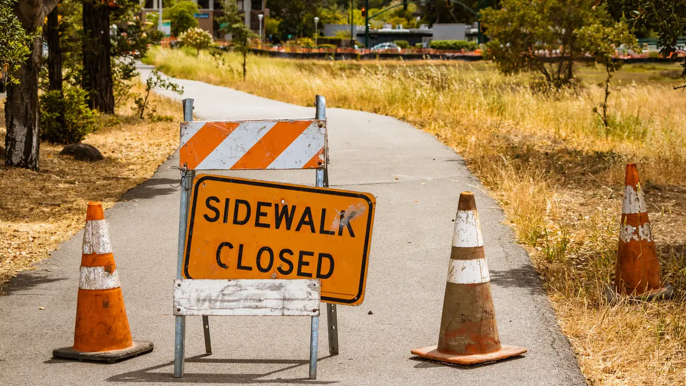 A paved walking path narrowed by a closed-sidewalk sign and orange traffic cones.